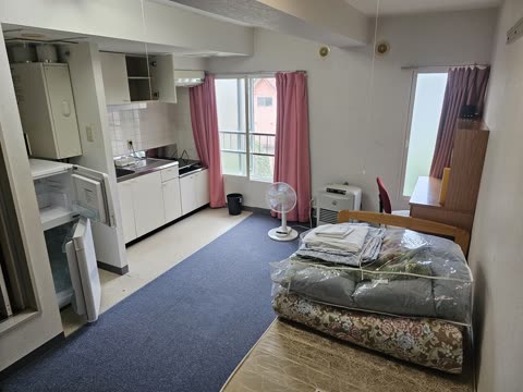 A studio apartment with a kitchenette along one wall and a bed with folded bedding in plastic bags.