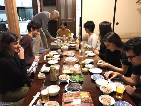 A group of people sits around a long, low table sharing a large meal of Japanese food inside a dining room.