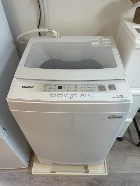 A white, top-loading Comfee washing machine with a Japanese control panel is installed against a wall on a wood-look floor.