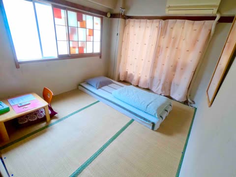 A simple bedroom with a tatami mat floor features a futon mattress, a low desk with a small chair, and a large window.