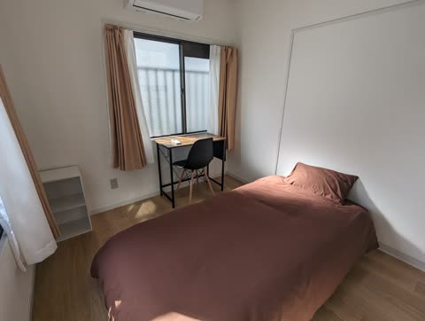 A bedroom with a bed made with brown linens, a small desk and black chair by a window, and white walls.