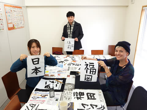 Three people around a table hold up papers with Japanese calligraphy characters in a brightly lit room.