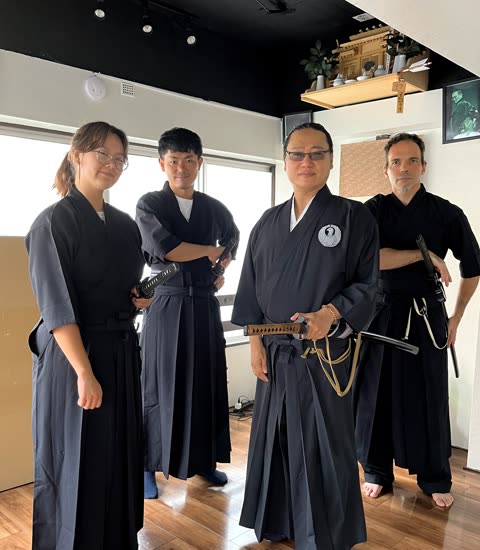 Four people in black martial arts attire stand indoors, holding swords.