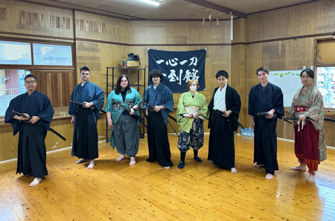 Seven people in traditional Japanese clothing holding swords, standing on a wooden floor.