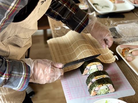 A person in an apron and gloves slices a sushi roll with a knife on a cutting mat at a wooden table.