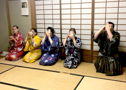 Five people in colorful kimonos kneel on a tatami mat floor, drinking from small bowls in front of a shoji screen.