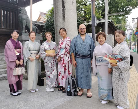 Seven people in kimonos pose outdoors near a Japanese shrine.