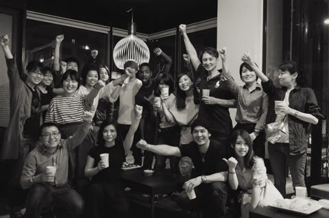 A large group of people stands and sits together indoors, smiling and raising their fists in the air.