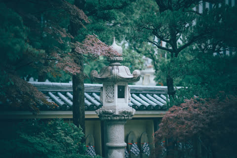 An ornate stone lantern stands in a garden surrounded by pine and maple trees in front of a tiled roof.