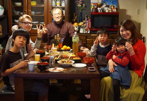 A group of adults and children sit at a dining table filled with food, looking at the camera and making peace signs.