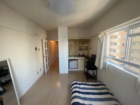 A compact studio apartment with a striped bed, a small kitchenette, and a window overlooking another building.