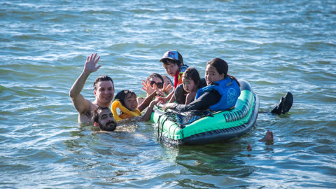 Group of people floating on an inflatable raft in water, one person waving.