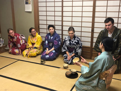 Woman in kimono demonstrates tea ceremony to group seated on tatami mats.