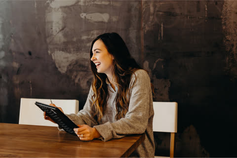 Woman smiles while writing on a tablet at a wooden table.