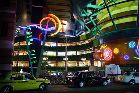 Person driving taxi in front of Canal City Hakata at night.