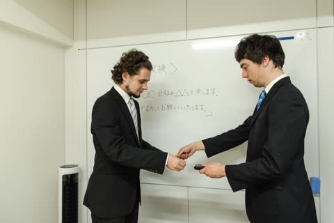 Two men in suits exchange cards in front of a whiteboard.