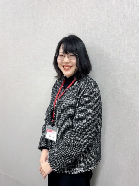 A woman with short black hair and glasses smiles while standing with her hands clasped in front of a gray wall.