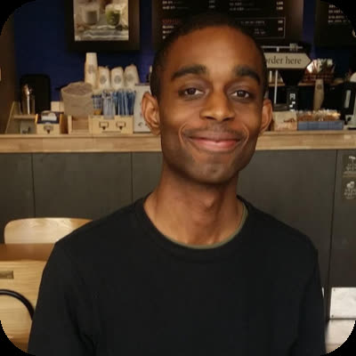 A man in a black shirt smiles at the camera while sitting inside a coffee shop.