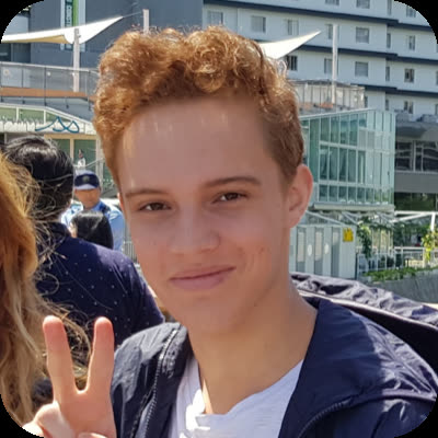 A young person with curly, reddish-brown hair holds up a peace sign while standing outdoors in front of a modern building.