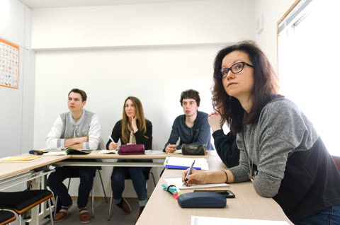 A woman with glasses writes in a notebook while looking up, seated with other people at a desk in a classroom.