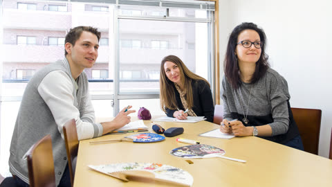 Three people sit at a wooden table with notebooks and pens in a room, looking toward the left.