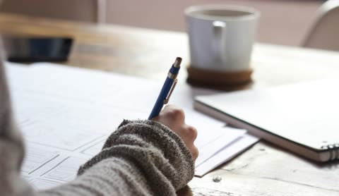 Person's hand writing on paper at a wooden desk.